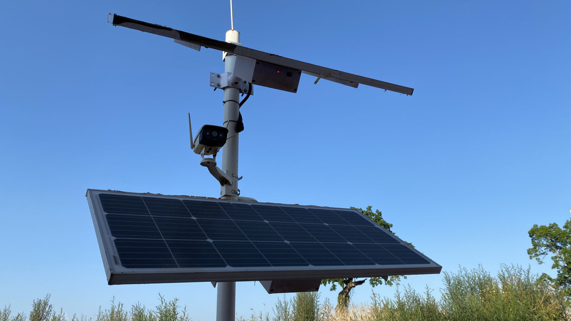 A solar panel and surveillance camera mounted on a pole, set against a clear blue sky with trees and bushes in the background.