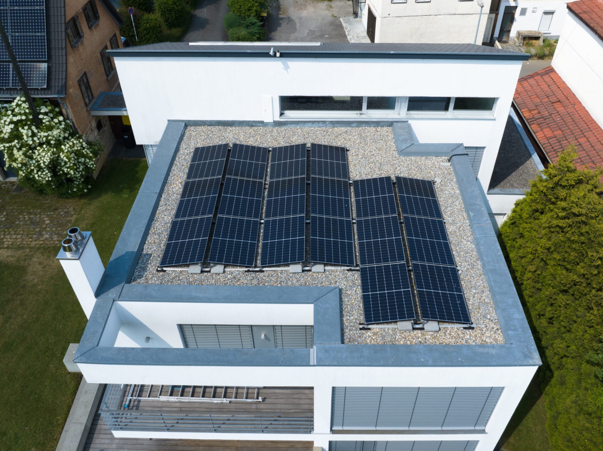 Modern home with flat rooftop solar panels, surrounded by greenery and neighboring houses, under a clear sky. Sustainable energy emphasis is evident.