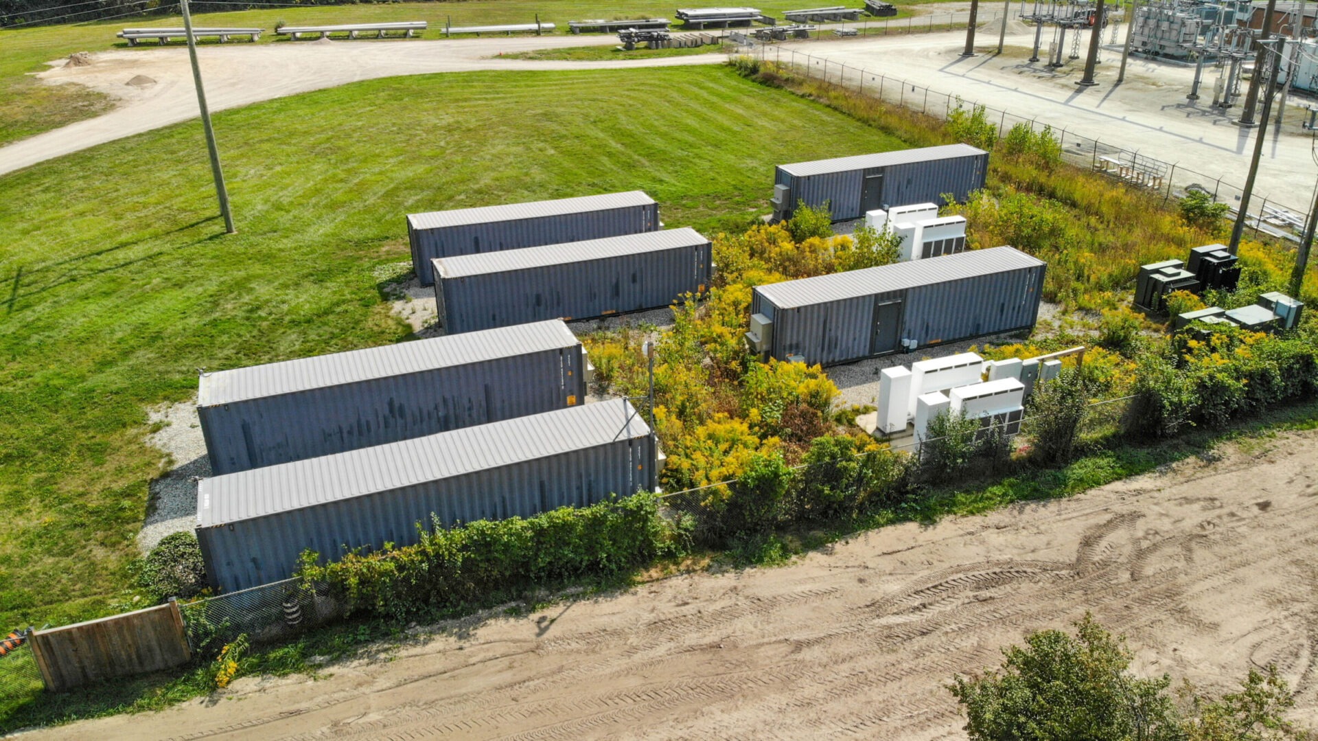 Aerial view of a grassy area with several shipping containers surrounded by trees and bushes near a power station. No people visible.